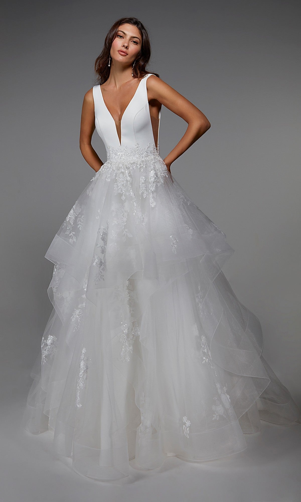 A woman wearing the Alyce 7040 long formal dress in ivory, featuring a sleeveless deep V-neck bodice and a layered tulle skirt adorned with floral lace.